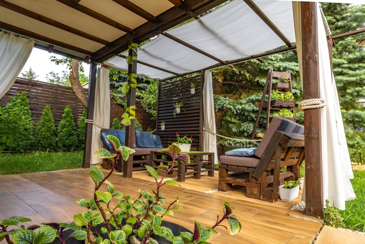 The image shows a cozy outdoor patio area with wooden furniture and a pergola. The seating includes cushioned chairs and a small table made from repurposed wood pallets. The pergola is adorned with hanging plants and climbing vines, providing a natural and inviting atmosphere. The patio is surrounded by a wooden fence and lush greenery, including trees and shrubs, which enhance the sense of privacy and tranquility. The floor is covered with wooden decking, and there are curtains on the pergola that can be drawn for additional shade or privacy.
