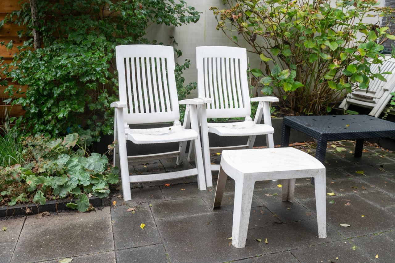 The image shows a pair of white plastic chairs and a matching white plastic table set on a patio. The chairs and table have a simple design, and they are placed next to some greenery, including bushes and plants, which add a natural element to the setting. The patio has a tiled floor, and there is another piece of outdoor furniture in the background, suggesting a casual and functional outdoor space. 