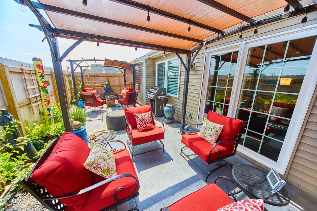 The image shows a vibrant and cozy outdoor patio space furnished with red cushioned chairs and a small table. The patio is covered by a pergola with string lights hanging from the frame, creating a warm and inviting atmosphere. The area also features a barbecue grill, various potted plants, and decorative elements, adding to the overall charm and functionality of the space. The patio is enclosed by a wooden fence, providing privacy and a sense of seclusion.