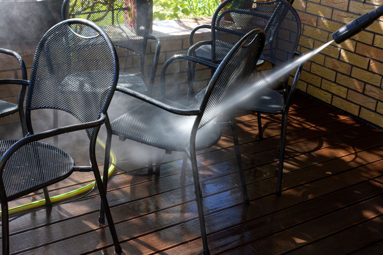 The image shows a person using a pressure washer to clean metal patio chairs. The spray of water is visible, effectively removing dirt and debris from the chairs. The chairs are made of metal with a mesh design and are placed on a wooden deck. The background includes a brick wall and some greenery, indicating an outdoor setting.