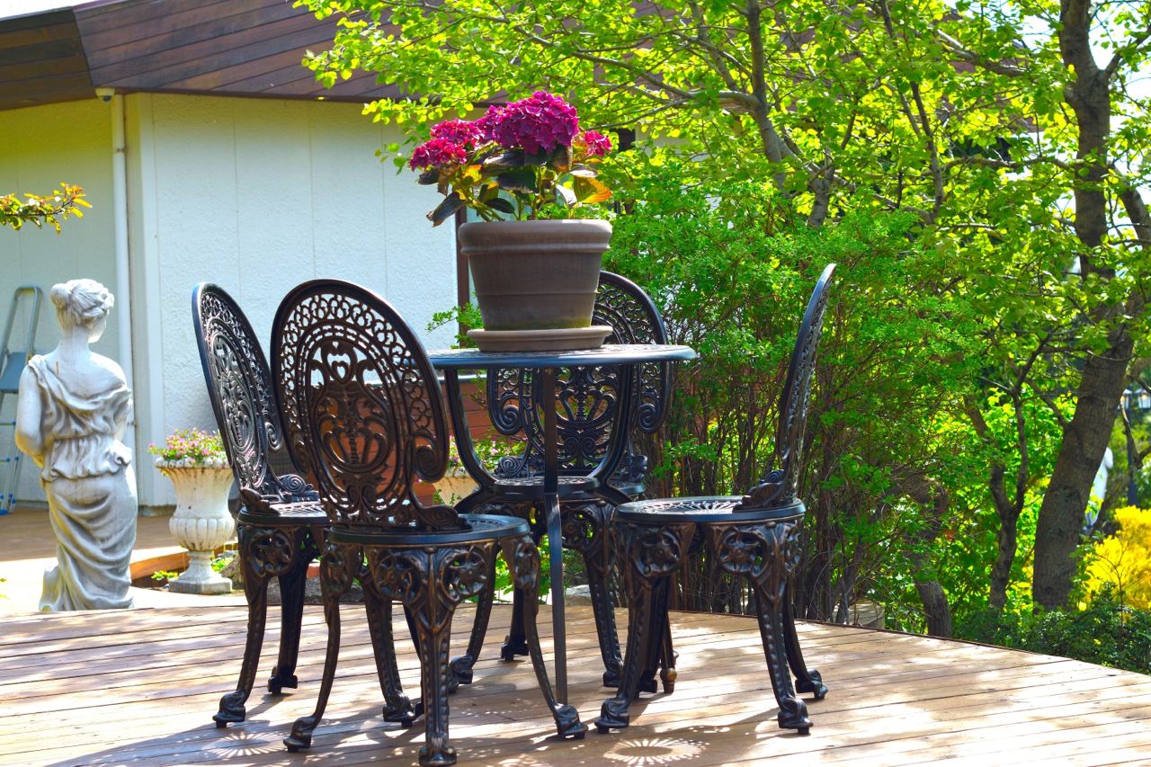 The image shows an elegant outdoor patio setting with a small round table and matching ornate chairs made of black wrought iron. On the table, there is a potted plant with vibrant pink flowers, adding a touch of color to the scene. The patio is surrounded by lush greenery, including bushes and trees, creating a peaceful and inviting atmosphere. In the background, a white statue adds a classical touch to the garden decor. 