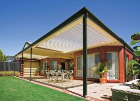 Spacious outdoor patio with a modern slatted pergola attached to a red-brick house, featuring a dining table and chairs, surrounded by potted plants and overlooking a green lawn under a clear blue sky.