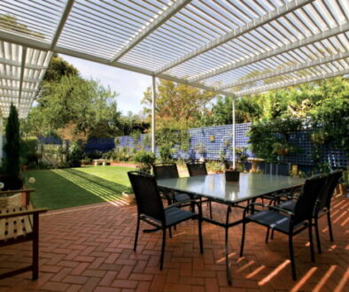 Open-roof Stratco Outback pergola attached to a house, shading a six-seat outdoor dining setting on brick paving