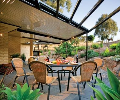 Outdoor dining area with a black-framed pergola, featuring a table set for a meal with chairs and lush greenery in the background. The space is surrounded by a landscaped garden with rocks and trees.