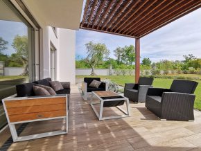 Modern outdoor seating area featuring black wicker chairs and sofas with wooden and metal frames under a pergola. The space has a wooden deck floor and overlooks a green garden with trees and a white wall in the background.